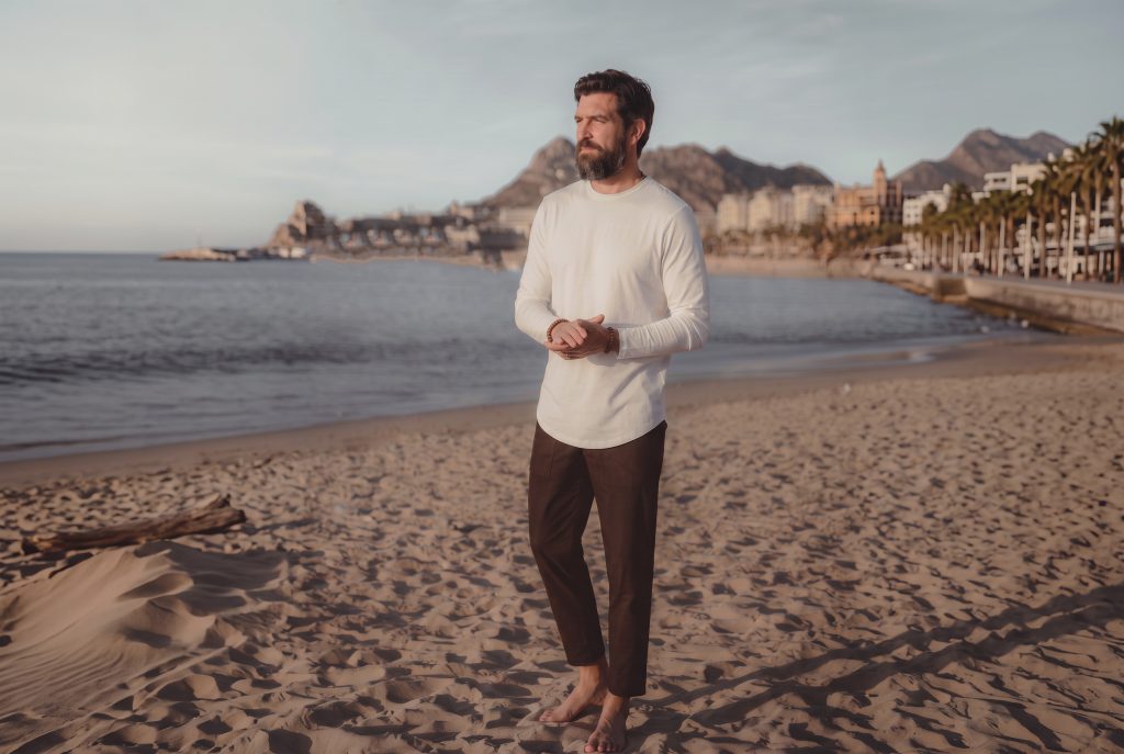 man on a beach in a long sleeve curved hem and relaxed brown pants